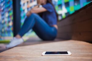 A smartphone lies on a wooden bench in the foreground, while a woman sits in the background with her knees up, looking distressed. The background shows colorful buildings, and the image conveys a sense of anxiety or emotional distress possibly related to digital harassment.